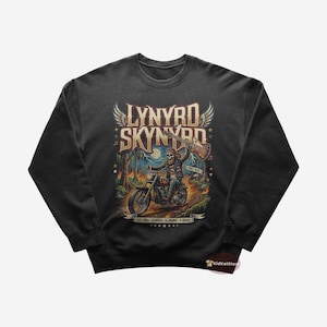 May include: Dark gray sweatshirt featuring a colorful graphic of a skeleton riding a motorcycle with a guitar. The design includes the band name "Lynyrd Skynyrd" and the text "Free Bird". The sweatshirt is a crew neck style.