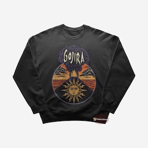 Op de afbeelding: Donkergrijze sweatshirt met een cirkelvormige afbeelding met een vulkaan, zon en tekst. Het woord "GOJIRA" staat in een gestileerd lettertype boven de vulkaan. De zon heeft een gezicht en stralen. De achtergrond toont een zonsondergang.