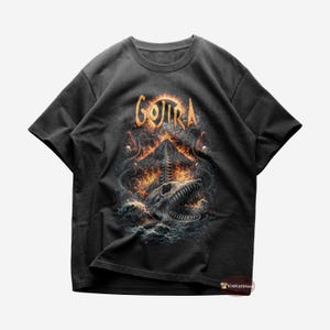 Op de afbeelding: Donkergrijs t-shirt met het woord "GOJIRA" in oranje, boven een gedetailleerde afbeelding. De afbeelding toont een schedel met scherpe tanden die uit het water oprijst, omringd door vlammen en tentakels. Het ontwerp heeft een heavy metal thema.