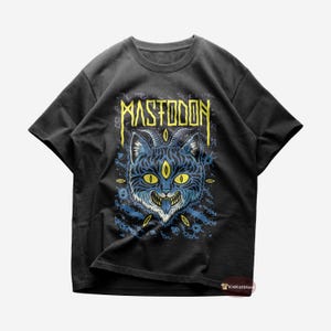 Puede incluir: Camiseta negra con un gráfico azul y amarillo de un gato con tres ojos y cuernos. El texto "MASTODON" está por encima del gato.
