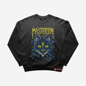 Puede incluir: Sudadera negra con un gráfico azul y amarillo de un gato con tres ojos y la palabra "MASTODON" impresa en amarillo.