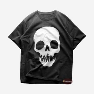 Può includere: T-shirt grigio scuro con grafica di teschio bianco e la scritta "GOJIRA" stampata sotto. Il design è una dichiarazione grafica audace. La maglietta è in stile girocollo classico.