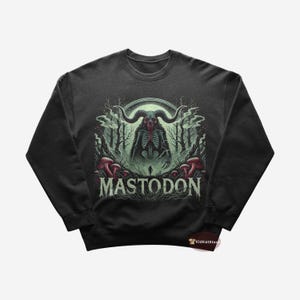 Puede incluir: Una sudadera gris oscuro con un estampado gráfico de una criatura esquelética en un entorno de bosque. El texto "MASTODON" está impreso debajo de la imagen.