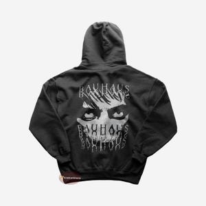 Puede incluir: Una sudadera con capucha gris oscuro con un estampado gráfico blanco de una cara y el texto "BAUHAUS" repetido varias veces.