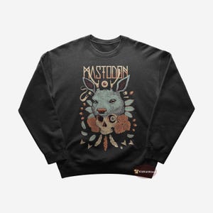 Puede incluir: Sudadera negra con un estampado gráfico de una calavera de ciervo con un ojo, rodeada de flores y plumas. El texto "MASTODON" está impreso sobre el gráfico.
