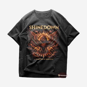May include: Black t-shirt featuring a detailed graphic of a phoenix with outstretched wings in shades of red, orange, and gold. The text "SHINEDOWN" is above the phoenix, and "A SYMPTOM OF SURVIVAL" is below.