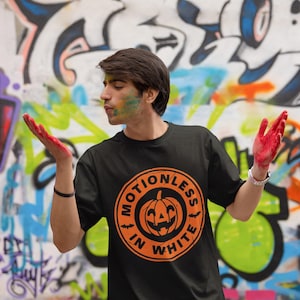 Może przedstawiać: Czarny t-shirt z pomarańczowym kołem graficznym przedstawiającym dynię z twarzą i tekstem "Motionless in White".