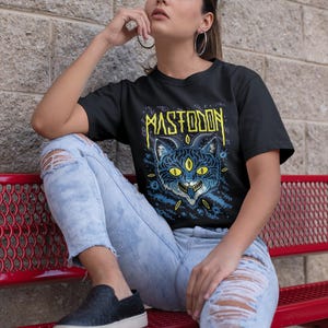 May include: A woman wearing a black t-shirt with a blue and yellow graphic of a three-eyed cat and the word "MASTODON" printed on it. She is wearing blue jeans and black slip-on shoes.