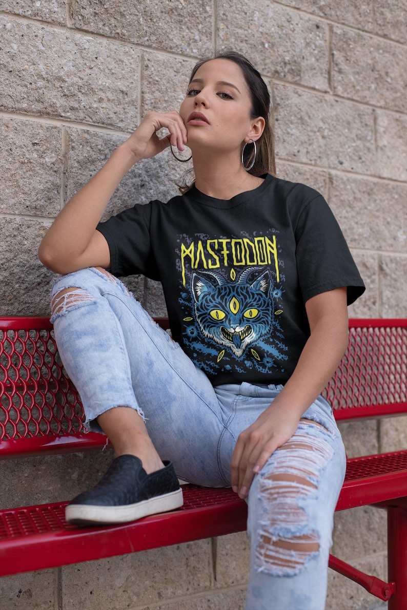 May include: A woman wearing a black t-shirt with a blue and yellow graphic of a three-eyed cat and the word "MASTODON" printed on it. She is wearing blue jeans and black slip-on shoes.
