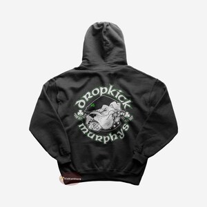 Peut inclure: Un sweat à capuche gris foncé avec un graphique blanc d'un bouledogue portant un chapeau et fumant un cigare. Le graphique est dans un cercle avec le texte "Dropkick Murphys" autour.