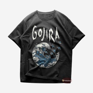 Op de afbeelding: Zwart T-shirt met een witte afbeelding van een golf en het woord "GOJIRA" erop gedrukt.