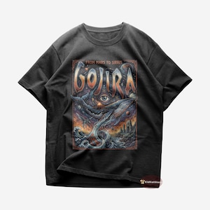 Op de afbeelding: Donkergrijs t-shirt met een kleurrijk grafisch ontwerp. Het ontwerp bevat het woord "GOJIRA" in grote letters, met een illustratie van een walvis, oog en oceaangolven. De tekst "FROM MARS TO SIRIUS" staat boven het woord "GOJIRA".
