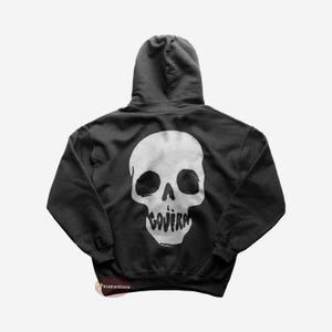 Puede incluir: Sudadera con capucha negra con un gráfico de calavera blanca en la espalda. La calavera tiene un contorno blanco y la palabra "GOJIRA" escrita dentro de la boca.