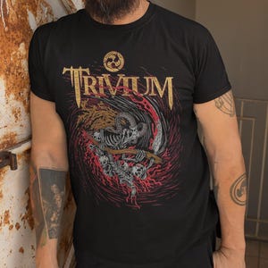 May include: Black t-shirt with a gold and red graphic of a skeleton holding a scythe in a swirling vortex. The text "TRIVIUM" is printed in gold above the graphic.