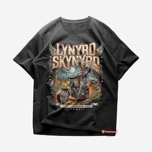 Puede incluir: Camiseta negra con un gráfico colorido de un esqueleto montando una motocicleta y tocando la guitarra. El diseño incluye el nombre de la banda "Lynyrd Skynyrd" y el texto "Free Bird". La camiseta también tiene el texto "Est. 1964 - Georgia - Alabama - Florida".