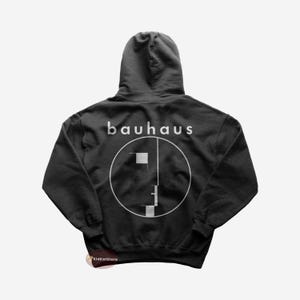 Puede incluir: Sudadera con capucha negra con un logotipo blanco de Bauhaus y un gráfico de cara estilizado en la parte posterior.