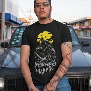 Może przedstawiać: Czarny t-shirt z grafiką z napisem "MOTIONLESS IN WHITE". Projekt zawiera żółte słońce, ptaki i postacie. Koszulka jest noszona przez osobę z tatuażami i okularami przeciwsłonecznymi.