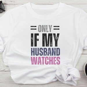 Puede incluir: Camiseta blanca con el texto "= ONLY = IF MY HUSBAND WATCHES" en una fuente desgastada. El texto es en gris, negro, azul y rosa. La camiseta está doblada sobre una superficie de madera clara.