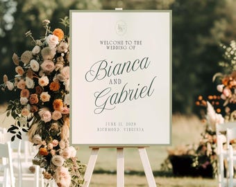 Puede incluir: Un caballete de madera blanco sostiene un letrero que dice "Welcome to the Wedding of Bianca and Gabriel" en una elegante escritura. El letrero, enmarcado en verde, está colocado sobre un césped, con arreglos florales y sillas blancas al fondo.