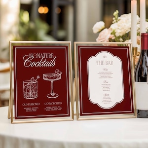Puede incluir: Dos letreros enmarcados en dorado sobre un mantel blanco. El letrero izquierdo dice "Signature Cocktails" y presenta ilustraciones de un Old Fashioned y un Cosmopolitan. El letrero derecho dice "The Bar" con una lista de bebidas.