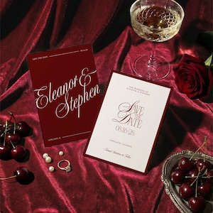 Op de afbeelding: Bordeaux trouwuitnodigingen met de namen "Eleanor & Stephen" en "Save the Date" op een rode fluwelen ondergrond. Een glas champagne, kersen, parels en een ring completeren de elegante sc&egrave;ne.