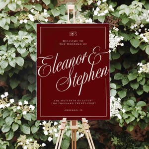 K&ouml;nnte beinhalten: Burgunderfarbenes Hochzeits-Willkommensschild auf einer Holzstaffelei. Das Schild zeigt eine elegante wei&szlig;e Schrift mit der Aufschrift &bdquo;Welcome to the Wedding of Eleanor & Stephen&ldquo;. Zus&auml;tzlicher Text enth&auml;lt Datum und Ort.