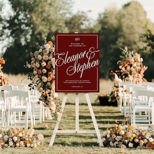 K&ouml;nnte beinhalten: Ein weinrotes Hochzeits-Willkommensschild auf einer wei&szlig;en Staffelei. Auf dem Schild steht "Willkommen zur Hochzeit von Eleanor & Stephen" mit Datum und Ort. Die Szene umfasst Blumengestecke und wei&szlig;e St&uuml;hle.