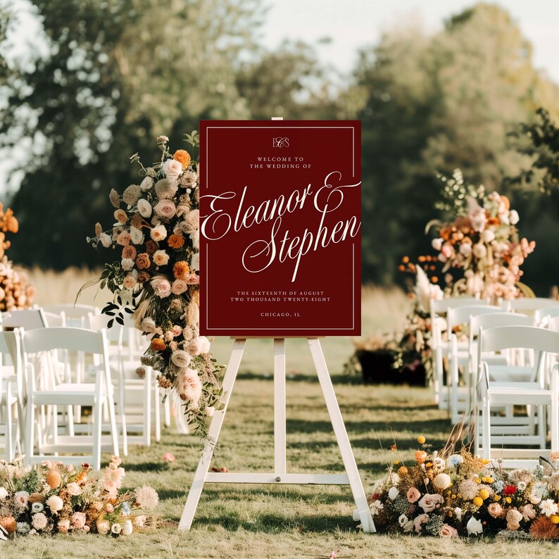K&ouml;nnte beinhalten: Ein weinrotes Hochzeits-Willkommensschild auf einer wei&szlig;en Staffelei. Auf dem Schild steht "Willkommen zur Hochzeit von Eleanor & Stephen" mit Datum und Ort. Die Szene umfasst Blumengestecke und wei&szlig;e St&uuml;hle.