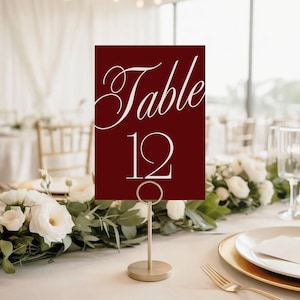 May include: A burgundy table number card with the word "Table" in elegant white script and the number "12" in a clean font. The card is held by a gold metal stand, set on a table with floral arrangements and place settings.