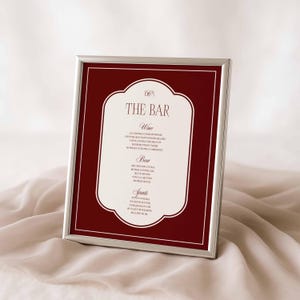 Puede incluir: Un men&uacute; de bar enmarcado con un marco plateado. El men&uacute; presenta un fondo burdeos con un borde blanco adornado. El texto "THE BAR" est&aacute; en la parte superior, seguido de listas de vinos, cervezas y licores. El men&uacute; se muestra sobre una tela suave de color crema.