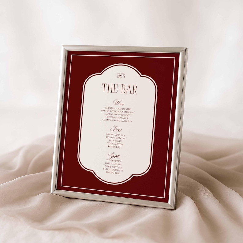 Puede incluir: Un men&uacute; de bar enmarcado con un marco plateado. El men&uacute; presenta un fondo burdeos con un borde blanco adornado. El texto "THE BAR" est&aacute; en la parte superior, seguido de listas de vinos, cervezas y licores. El men&uacute; se muestra sobre una tela suave de color crema.