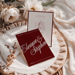 Op de afbeelding: Bordeauxrode en cr&egrave;mekleurige huwelijksuitnodigingen met elegant schrift. De tekst luidt "Save the Date" en "Eleanor & Stephen". De uitnodigingen zijn gerangschikt op een geweven dienblad met witte bloemen en stof.