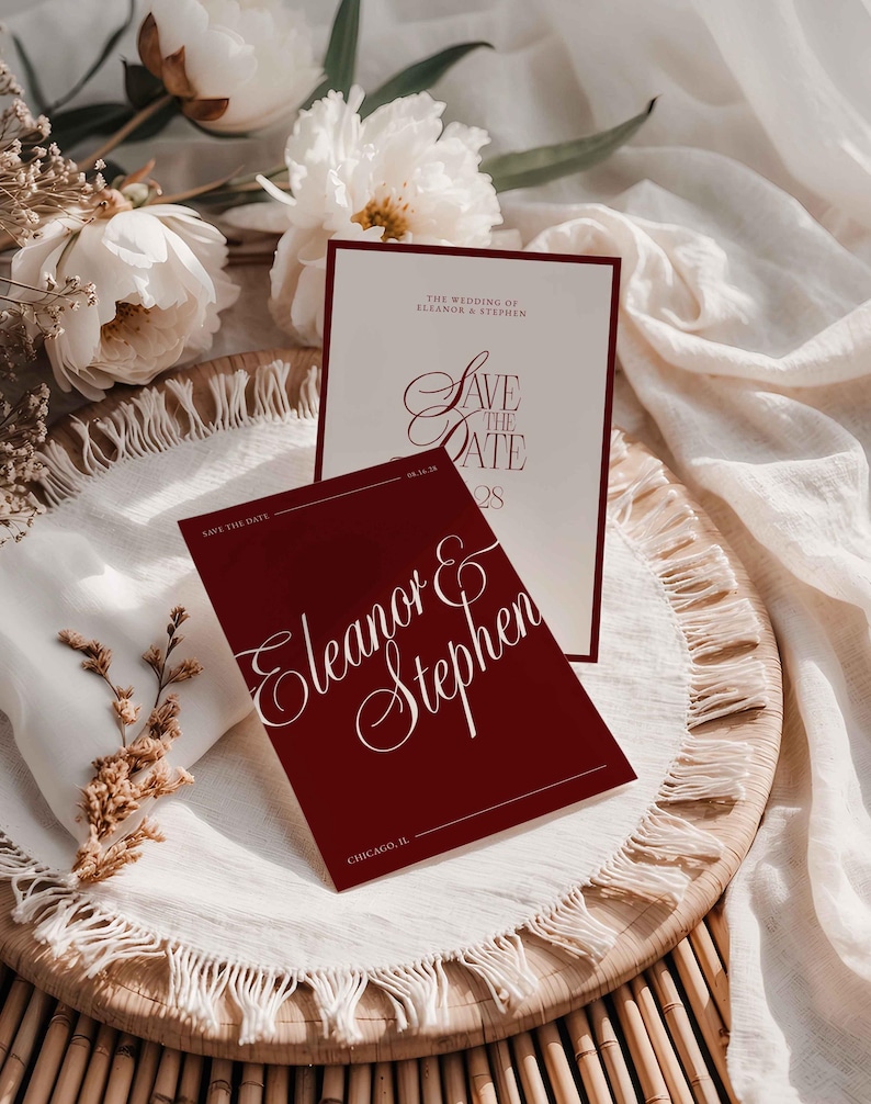 Op de afbeelding: Bordeauxrode en cr&egrave;mekleurige huwelijksuitnodigingen met elegant schrift. De tekst luidt "Save the Date" en "Eleanor & Stephen". De uitnodigingen zijn gerangschikt op een geweven dienblad met witte bloemen en stof.