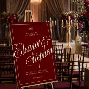 K&ouml;nnte beinhalten: Ein weinrotes Hochzeits-Willkommensschild auf einer goldenen Staffelei. Das Schild tr&auml;gt den wei&szlig;en Text "Willkommen zur Hochzeit von Eleanor & Stephen" und das Datum. Der Hintergrund umfasst St&uuml;hle, Blumen und Kronleuchter.