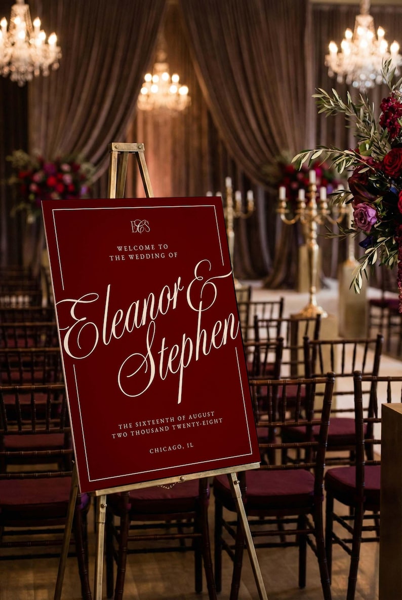 K&ouml;nnte beinhalten: Ein weinrotes Hochzeits-Willkommensschild auf einer goldenen Staffelei. Das Schild tr&auml;gt den wei&szlig;en Text "Willkommen zur Hochzeit von Eleanor & Stephen" und das Datum. Der Hintergrund umfasst St&uuml;hle, Blumen und Kronleuchter.
