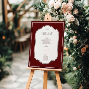 Puede incluir: Un letrero burdeos sobre un caballete de madera con la frase "The Bar" en una fuente elegante. El letrero enumera vinos, cervezas y licores. El fondo presenta arreglos florales, ideal para una boda.
