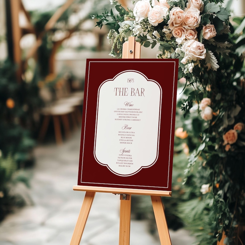 Puede incluir: Un letrero burdeos sobre un caballete de madera con la frase "The Bar" en una fuente elegante. El letrero enumera vinos, cervezas y licores. El fondo presenta arreglos florales, ideal para una boda.