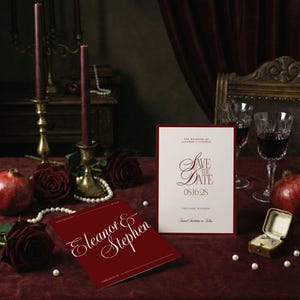Op de afbeelding: Een romantische sc&egrave;ne met trouwbriefpapier. Twee kaarten met elegant schrift met de tekst "Eleanor & Stephen" en "Save the Date" worden getoond. De sc&egrave;ne omvat rozen, parels, kaarsen en een ringdoosje, allemaal op een bordeauxrode fluwelen ondergrond.