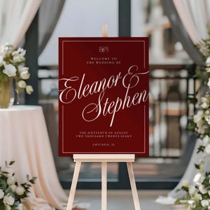 K&ouml;nnte beinhalten: Ein weinrotes Hochzeits-Willkommensschild auf einer Holzstaffelei. Das Schild zeigt eine elegante wei&szlig;e Schrift mit der Aufschrift "Willkommen zur Hochzeit von Eleanor & Stephen" sowie das Datum "Der sechzehnte August zweitausendachtundzwanzig" und den Ort "Chicago, IL".