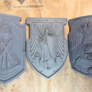 May include: Three grey, shield-shaped decorative pieces with detailed relief designs. The shields feature various emblems, including a skull, a figure with a sword, and crossed weapons, suggesting a fantasy or historical theme.