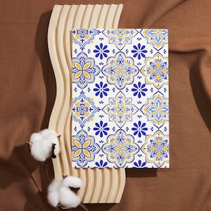 May include: A patterned notebook with a white background and blue and yellow floral designs. The notebook is displayed on a light wood decorative piece with a brown background. Two cotton flowers are in the foreground.