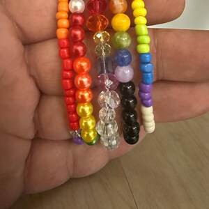 Puede incluir: Una colección de pulseras de cuentas coloridas, cada una con un esquema de color diferente. Las pulseras incluyen cuentas naranjas, rojas, amarillas, verdes, azules y moradas, algunas incorporando cuentas transparentes e iridiscentes. Las cuentas están ensartadas en un cordón fino y flexible.