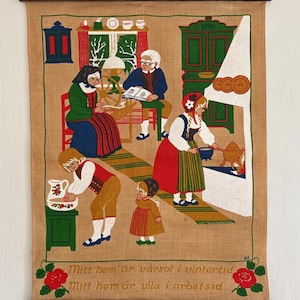 May include: A vintage tapestry depicting a family gathered in a cosy home. The scene features a fireplace, a spinning wheel, and a table with a book. The tapestry is made of burlap and has a warm, rustic feel. The text on the tapestry reads "Mitt hem ar varsol i vintertid Mitt hem ar vila i arbetsid."
