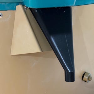 May include: A close-up of a black plastic funnel attached to a teal blue container. The funnel is attached to a beige surface.