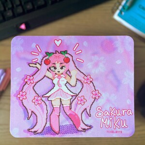 May include: A pink mousepad featuring a pixelated illustration of Sakura Miku, a character with long pink hair, a white dress, and pink boots. The mousepad has the text "Sakura Miku" and "TIXEL.BYTE" printed on it. The background is a light pink with floral patterns.