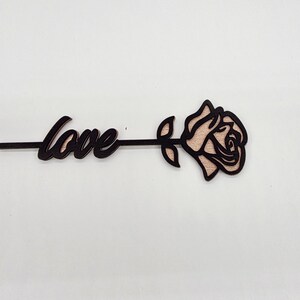 以下が含まれることがあります： 木製の装飾品で、筆記体の「love」の文字とバラが組み合わされています。 バラは薄いピンク色の中心部と黒いアウトラインを持っています。 全体は白い背景に配置されています。
