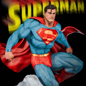 Op de afbeelding: Een Superman-beeld in een dynamische pose, gekleed in zijn iconische blauwe pak, rode broek en cape. De figuur heeft een gedetailleerde sculptuur, met het Superman-logo op de borst. De achtergrond toont het woord "SUPERMAN" in geel.
