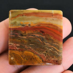 Puede incluir: Piedra cuadrada con superficie pulida. La piedra presenta capas de color, incluyendo tonos de amarillo, naranja, rojo y marrón. El patrón se asemeja a un paisaje o diseño abstracto. La piedra se sostiene en una mano.