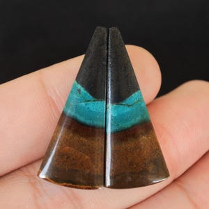 以下が含まれることがあります： 研磨された三角形の石が2つ。手で持たれており、黒、ターコイズ、茶色のグラデーションカラーです。これらの石は、おそらくジュエリーや装飾目的で使用されます。