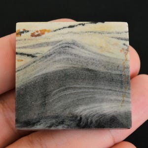 May include: A square stone with a natural pattern of swirling grey, black, and cream colours. The stone has a polished surface and a unique, organic design. The colours include hints of orange and brown.